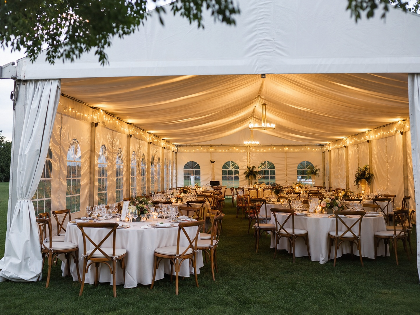 Interior de carpa modular blanca decorada para boda con telas drapeadas, lámparas de araña y mesas redondas iluminadas al atardecer